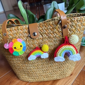 Peut inclure: Un sac fourre-tout en paille tressée avec des anses en cuir marron et trois breloques au crochet. Les breloques comprennent une fleur souriante et deux arcs-en-ciel avec des nuages blancs duveteux. Les breloques sont fixées au sac avec des anneaux métalliques dorés.