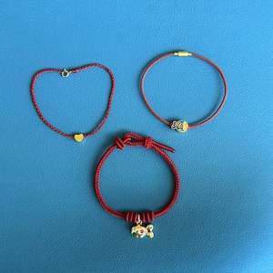 Op de afbeelding: Drie rode touwarmbanden met goudkleurige bedels. Eén armband heeft een hartbedel, een andere een visbedel en de derde een bedel met een bloemmotief. De armbanden worden op een blauw oppervlak weergegeven.