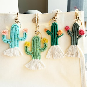 Pode incluir: Quatro chaveiros de cacto feitos à mão em tons de azul-turquesa, verde e cinza. Cada cacto tem uma base de borla branca, ferragens douradas e flores de pompom coloridas. Os chaveiros são exibidos em um fundo neutro.