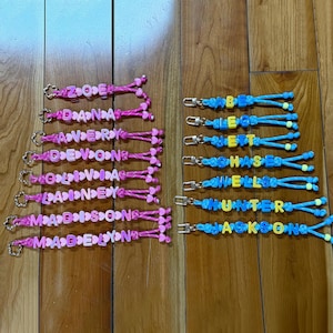 May include: A collection of personalised keychains featuring colourful plastic letter beads and heart-shaped spacers on braided cords. The left column displays pink cords with names like Zoe, Dana, Avery, Devon, Olivia, Lainey, Madison, and Madelyn. The right column features blue cords with names like Abe, Leo, Jett, Chase, Wells, Hunter, and Jackson, each attached to a silver metal lobster clasp.