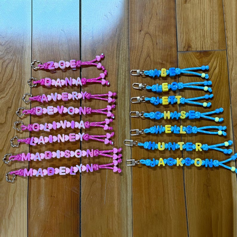 May include: A collection of personalised keychains featuring colourful plastic letter beads and heart-shaped spacers on braided cords. The left column displays pink cords with names like Zoe, Dana, Avery, Devon, Olivia, Lainey, Madison, and Madelyn. The right column features blue cords with names like Abe, Leo, Jett, Chase, Wells, Hunter, and Jackson, each attached to a silver metal lobster clasp.