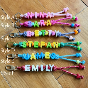 May include: Six keychains labelled Style A through F, each featuring colourful plastic letter beads spelling names like NINA, SARA, BETTY, STEFAN, JAMES, and EMILY, attached to metal lobster clasps with various coloured nylon cords and heart beads.