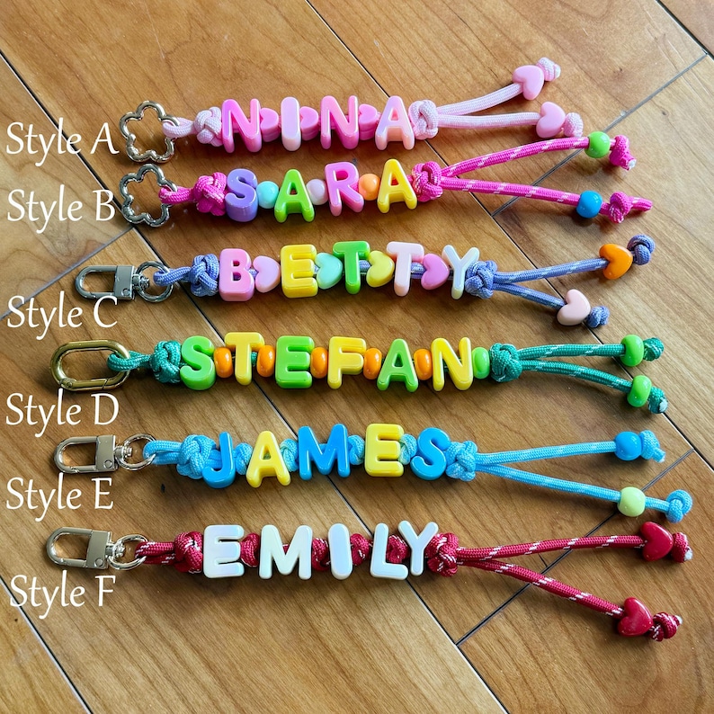 May include: Six keychains labelled Style A through F, each featuring colourful plastic letter beads spelling names like NINA, SARA, BETTY, STEFAN, JAMES, and EMILY, attached to metal lobster clasps with various coloured nylon cords and heart beads.