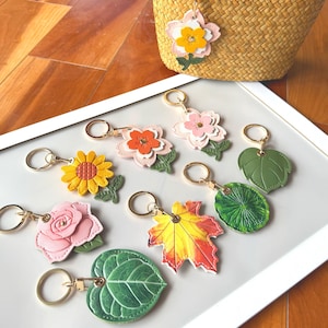 Könnte beinhalten: Eine Sammlung bunter Schlüsselanhänger in Blumen- und Blattform. Die Schlüsselanhänger zeigen Designs wie eine Sonnenblume, eine Rose, ein Ahornblatt und verschiedene Blumenformen, jeweils mit einem goldfarbenen Schlüsselring. Die Schlüsselanhänger sind auf einem weißen Tablett angeordnet.