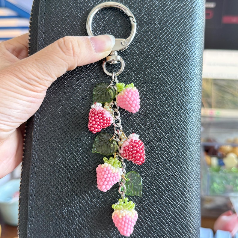 Pu&ograve; includere: Un portachiavi a forma di fragole di perline, con un anello color argento. Le fragole sono rosa e rosse, con cime e foglie verdi. Il portachiavi &egrave; attaccato a un portafoglio in pelle nera.