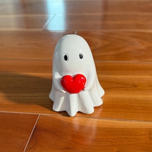 Puede incluir: Una figura de fantasma blanco sosteniendo un coraz&oacute;n rojo. El fantasma tiene ojos negros y una forma drapeada y fluida. El coraz&oacute;n es liso y brillante, contrastando con el acabado mate del fantasma.