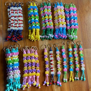 May include: A collection of keychains featuring colorful plastic letter beads spelling the word ELEVATE. Each keychain is constructed with a metal ring, various colored cord, and small accent beads, with some designs ending in heart-shaped charms.