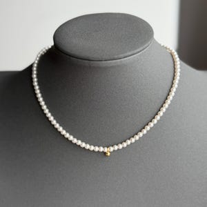 Peut inclure: Collier de perles d&eacute;licat avec de petites perles blanches rondes. Un petit charme dor&eacute; est suspendu au centre. Le collier est pr&eacute;sent&eacute; sur une t&ecirc;te de mannequin grise, mettant en valeur son design &eacute;l&eacute;gant.