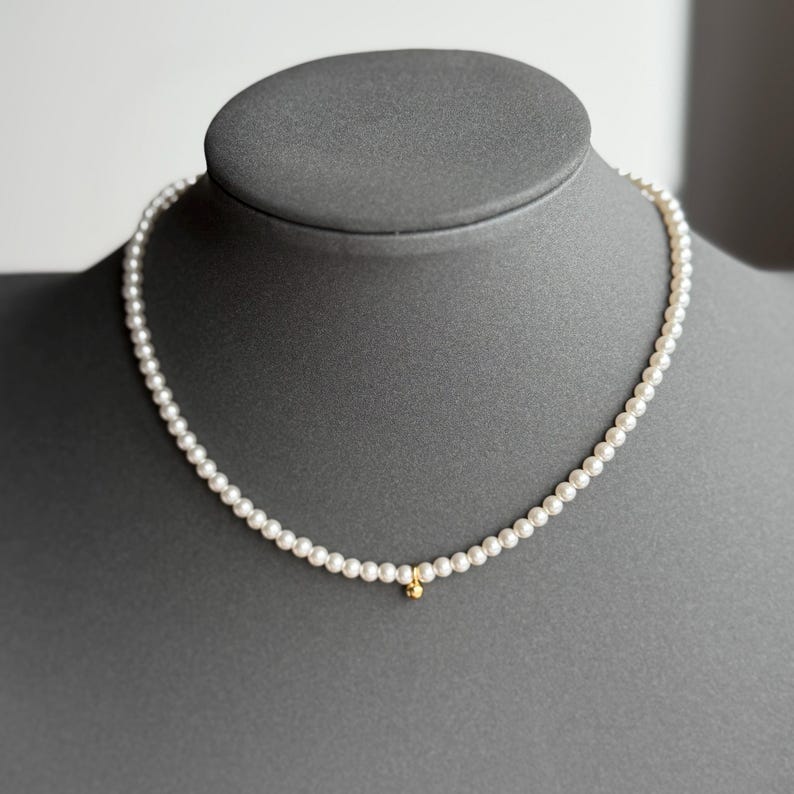 Peut inclure: Collier de perles d&eacute;licat avec de petites perles blanches rondes. Un petit charme dor&eacute; est suspendu au centre. Le collier est pr&eacute;sent&eacute; sur une t&ecirc;te de mannequin grise, mettant en valeur son design &eacute;l&eacute;gant.