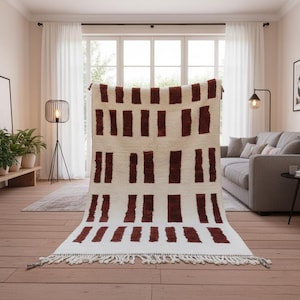 May include: A rectangular, cream-colored rug with a repeating pattern of vertical, dark red rectangles. The rug has a fringe along the bottom edge. The rug is displayed in a well-lit room with a hardwood floor and a large window.