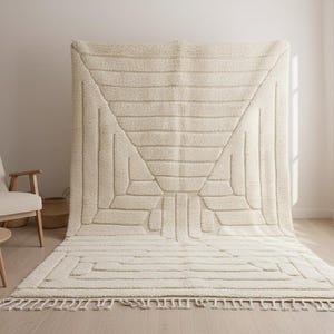 Puede incluir: Una alfombra grande de color crema con un diseño geométrico. La alfombra presenta un triángulo central y formas rectangulares, con una franja en el borde inferior. La alfombra se exhibe en una habitación con una silla y una mesa pequeña.
