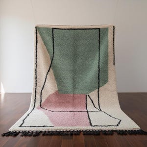 May include: A modern, abstract rug with a geometric design. The rug features a large green rectangle, a pink shape, and a cream-colored background, all outlined in black. Black tassels line the bottom edge.