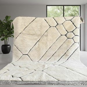 May include: A large, off-white rug with a geometric pattern of black lines and shapes. The rug has a thick, textured pile and a fringed edge with black and white stripes. A potted plant is in the background.