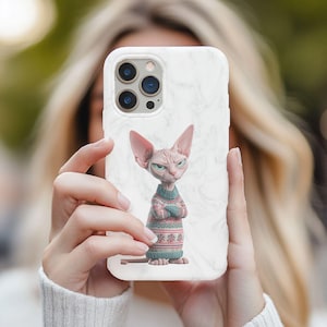 May include: White marble phone case featuring a Sphynx cat wearing a sweater. The cat has a grumpy expression with crossed arms. The sweater is blue, pink, and white with a snowflake pattern. The case has a triple-lens camera.