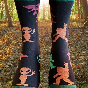 May include: Black socks with green cuffs and toes, patterned with orange aliens, pink UFOs, and green and orange Bigfoot figures. The socks are knee-high and are set against a blurred forest background.