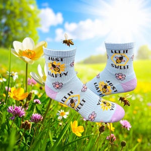 May include: A pair of light blue socks with a bee and flower design. The socks have the words "Just Happy You" and "Let It Sweet" printed on them. The socks have a pink heel and toe. The background is a sunny meadow with flowers and bees.