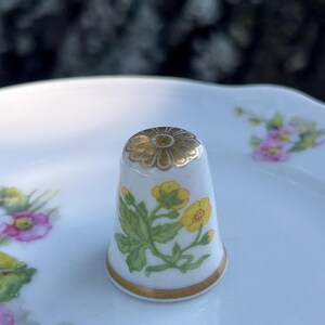 May include: A white porcelain thimble featuring a hand-painted yellow flower design with green leaves on the side and a gold-painted floral pattern on the top. The base of the thimble is finished with a thin gold band around the bottom edge.