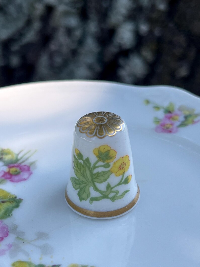 May include: A white porcelain thimble featuring a hand-painted yellow flower design with green leaves on the side and a gold-painted floral pattern on the top. The base of the thimble is finished with a thin gold band around the bottom edge.