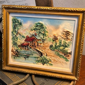 May include: A framed, three-dimensional landscape painting. The artwork depicts a rural scene with a house, watermill, bridge, and trees. The frame is gold-colored with ornate detailing. The painting's colors include shades of blue, green, brown, and red.