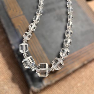 May include: A clear crystal necklace with faceted beads of varying sizes. The necklace features a central, larger geometric bead, with smaller, square-cut beads leading to the clasp. The necklace is displayed on a dark, textured surface.