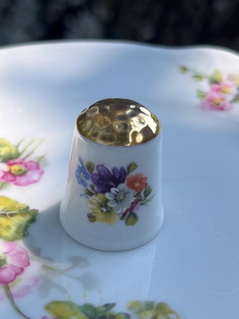 May include: A white porcelain thimble featuring a gold-toned, hammered metal top. The body of the thimble is decorated with a colourful floral bouquet print consisting of purple, yellow, orange, and blue flowers with green leaves.