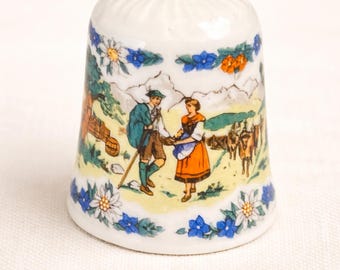 May include: A white ceramic thimble featuring a colourful scene of a couple in traditional dress, set against a mountain landscape. The thimble is decorated with blue and white floral patterns.
