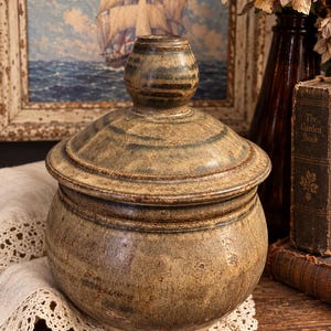 May include: A round, lidded ceramic jar with a mottled, earthy brown and muted green glaze. The jar features a wide, bulbous body and a matching lid with a prominent, rounded knob handle at the top. The surface has a textured, weathered appearance.