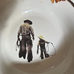 Puede incluir: Cuenco de cerámica blanca con diseño de vaqueros. La imagen muestra dos vaqueros, un adulto y un niño, con cuerdas. El cuenco tiene un borde ligeramente festoneado y grietas visibles.