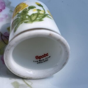 May include: A white fine bone china thimble featuring a yellow floral pattern. The interior base is marked with the red text Spode, Fine Bone China, and ENGLAND. The thimble has a smooth, rounded top and a flared base rim.