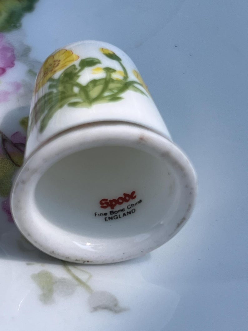 May include: A white fine bone china thimble featuring a yellow floral pattern. The interior base is marked with the red text Spode, Fine Bone China, and ENGLAND. The thimble has a smooth, rounded top and a flared base rim.