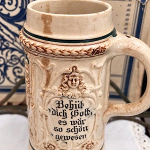 May include: A beige ceramic beer stein with a handle, featuring a relief-molded face and the German text "Behüt dich Gott, es wär so schön gewesen" in black lettering. The vessel has a glazed finish with visible crazing and dark green accents.