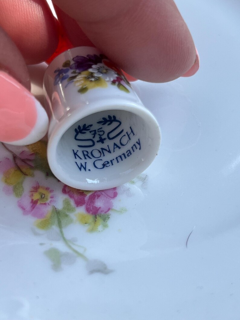 May include: A white porcelain thimble featuring a colourful floral pattern of purple, yellow, and pink flowers. The interior base is marked with blue text reading 75 S+C KRONACH W. Germany, indicating the origin and manufacturer of the item.