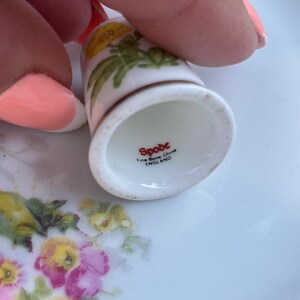 May include: A white fine bone china thimble featuring a yellow floral pattern. The interior base is stamped with the red text Spode, Fine Bone China, and ENGLAND. The thimble has a smooth, glossy finish and a flared rim at the bottom edge.