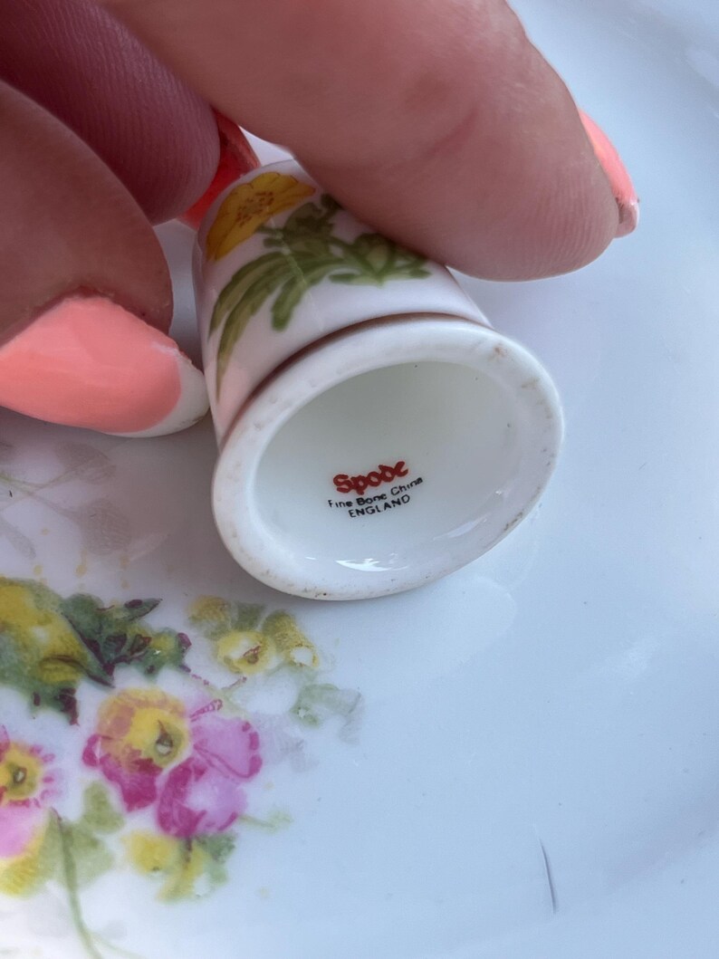 May include: A white fine bone china thimble featuring a yellow floral pattern. The interior base is stamped with the red text Spode, Fine Bone China, and ENGLAND. The thimble has a smooth, glossy finish and a flared rim at the bottom edge.