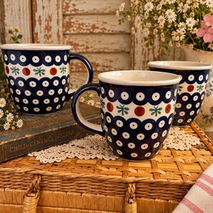 May include: Three ceramic mugs featuring a dark blue background with a repeating pattern of white circles, red dots, and green star-like shapes. The mugs have a white interior and a sturdy handle, showcasing a consistent folk-inspired design.