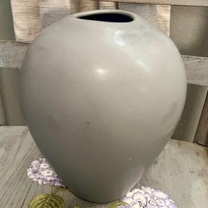 Op de afbeelding: Een grote, grijze keramische vaas met een afgeronde body en een smalle opening. De vaas heeft een gladde, glanzende afwerking en een donker interieur. De vaas is een decoratief item, geschikt voor het uitstallen van bloemen of als een op zichzelf staand accentstuk.