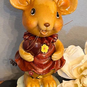 May include: A ceramic mouse figurine with large ears, holding a heart-shaped object. The mouse wears a red dress with a ruffled collar and a yellow flower. The figurine is on a light blue base.