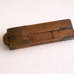 May include: A vintage wooden protractor with brass hinges and accents. The tool is rectangular when closed, with a circular hinge on one end. The wood has a warm brown tone, and the brass has a tarnished appearance. A classic woodworking tool.