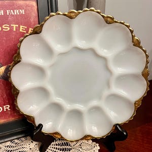 Peut inclure: Une assiette à œufs blanche et festonnée avec une bordure dorée. L'assiette comporte 12 compartiments individuels pour les œufs et une zone de service centrale. L'assiette est présentée sur un support noir, avec une image encadrée rouge et noire en arrière-plan.