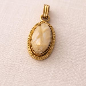 May include: Oval pendant with a carved cameo design in a gold-tone setting. The cameo features a light beige profile. The pendant has a twisted rope-like border and a decorative bail.