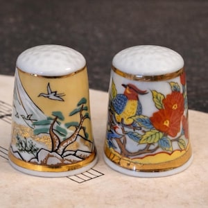 May include: Two decorative porcelain thimbles. One features a mountain landscape with a flying bird, the other a bird perched on a flowering branch. Both have gold trim and white tops.