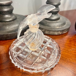 Puede incluir: Un frasco de perfume de vidrio transparente con un tapón en forma de pájaro, apoyado sobre una base de vidrio con diseño de diamantes. La botella tiene un diseño en espiral y el pájaro está representado en vuelo. La base tiene un borde festoneado.