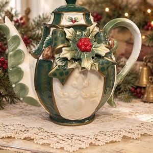 May include: A ceramic teapot featuring a cream-colored body with embossed scrollwork, accented by deep green panels and gold trim. The lid and spout are decorated with green leaves, while the front displays a sculpted holly and bow motif.