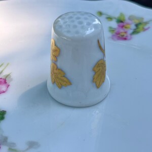 Peut inclure: Un d&eacute; &agrave; coudre en porcelaine blanche dot&eacute; d'un sommet alv&eacute;ol&eacute; et de motifs de feuilles dor&eacute;es peints sur les c&ocirc;t&eacute;s. Le d&eacute; pr&eacute;sente une finition lisse et brillante et est plac&eacute; &agrave; la verticale, mettant en valeur les d&eacute;tails du feuillage dor&eacute;.