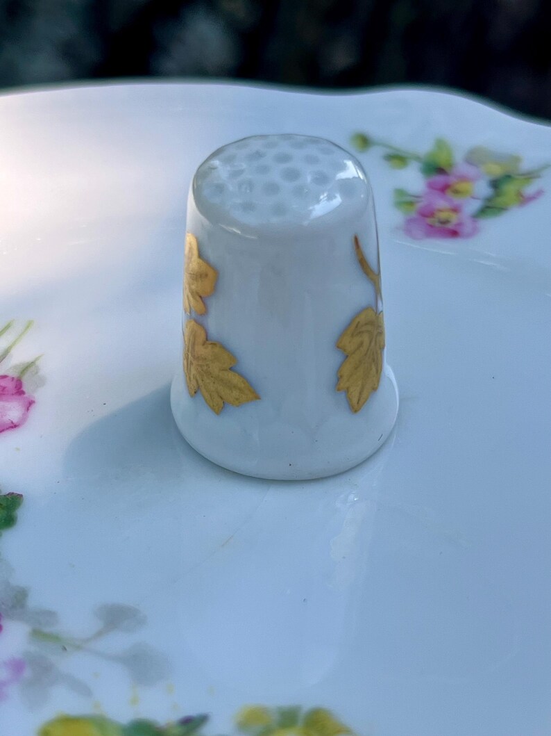 Peut inclure: Un d&eacute; &agrave; coudre en porcelaine blanche dot&eacute; d'un sommet alv&eacute;ol&eacute; et de motifs de feuilles dor&eacute;es peints sur les c&ocirc;t&eacute;s. Le d&eacute; pr&eacute;sente une finition lisse et brillante et est plac&eacute; &agrave; la verticale, mettant en valeur les d&eacute;tails du feuillage dor&eacute;.