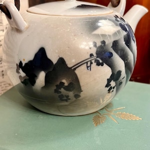 Puede incluir: Una tetera de cerámica blanca con un diseño de paisaje negro y azul. La tetera tiene un cuerpo redondeado, un asa pequeña y una tapa con un pequeño pomo. La tetera está sobre un libro verde claro.