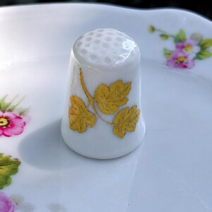 Peut inclure: Un d&eacute; &agrave; coudre en porcelaine blanche orn&eacute; d'un motif de feuilles dor&eacute;es sur le c&ocirc;t&eacute; et d'un sommet alv&eacute;ol&eacute;. Le d&eacute; est pos&eacute; sur une surface en c&eacute;ramique blanche d&eacute;cor&eacute;e de petits motifs floraux roses et verts.