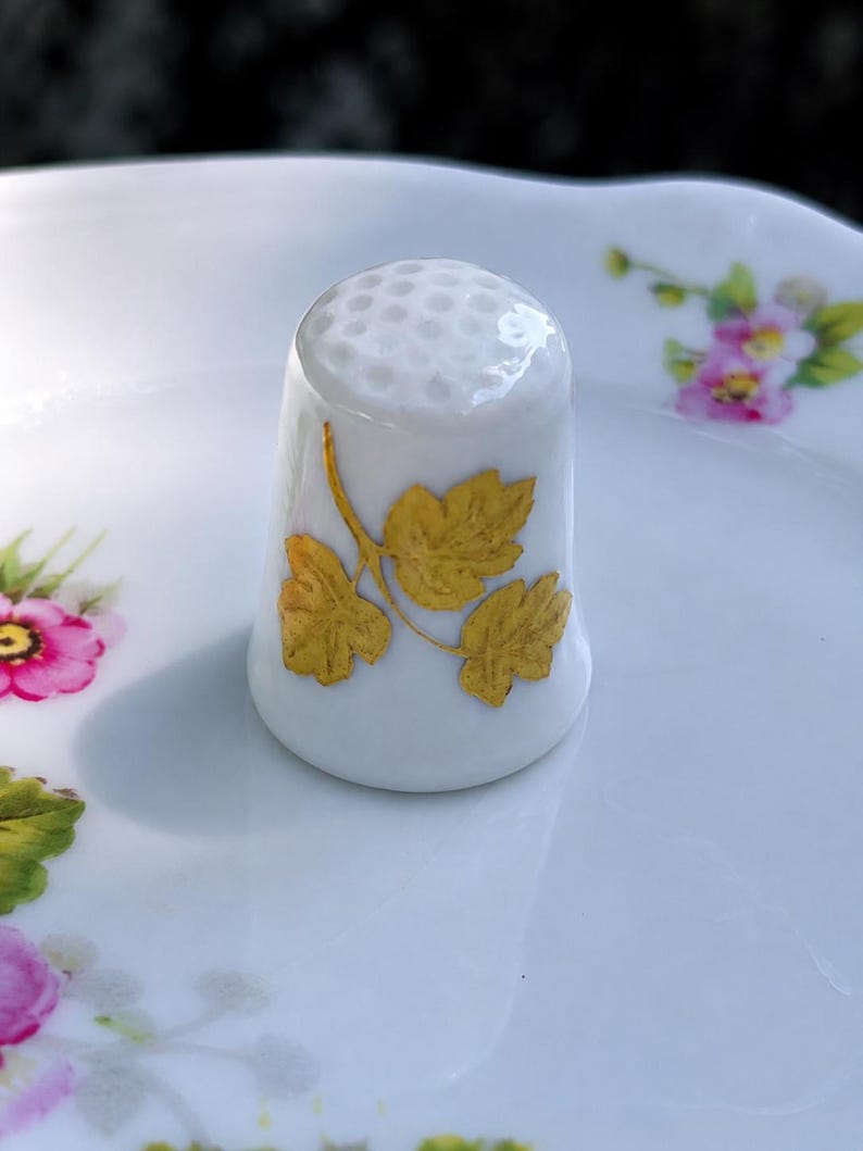 Peut inclure: Un d&eacute; &agrave; coudre en porcelaine blanche orn&eacute; d'un motif de feuilles dor&eacute;es sur le c&ocirc;t&eacute; et d'un sommet alv&eacute;ol&eacute;. Le d&eacute; est pos&eacute; sur une surface en c&eacute;ramique blanche d&eacute;cor&eacute;e de petits motifs floraux roses et verts.