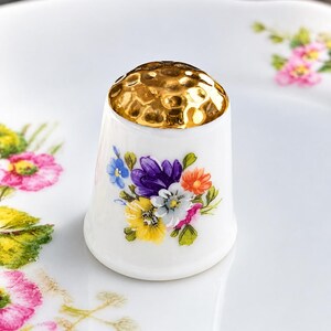 May include: A white ceramic thimble featuring a gold-toned, dimpled metal top and a colourful floral bouquet design on the side, showcasing purple, yellow, blue, orange, and white flowers with green leaves against a smooth, glossy finish.