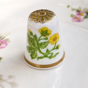 May include: A white porcelain thimble featuring a hand-painted yellow buttercup flower design with green leaves. The top of the thimble is decorated with a gold-colored floral pattern, and the base is finished with a thin gold-colored band.